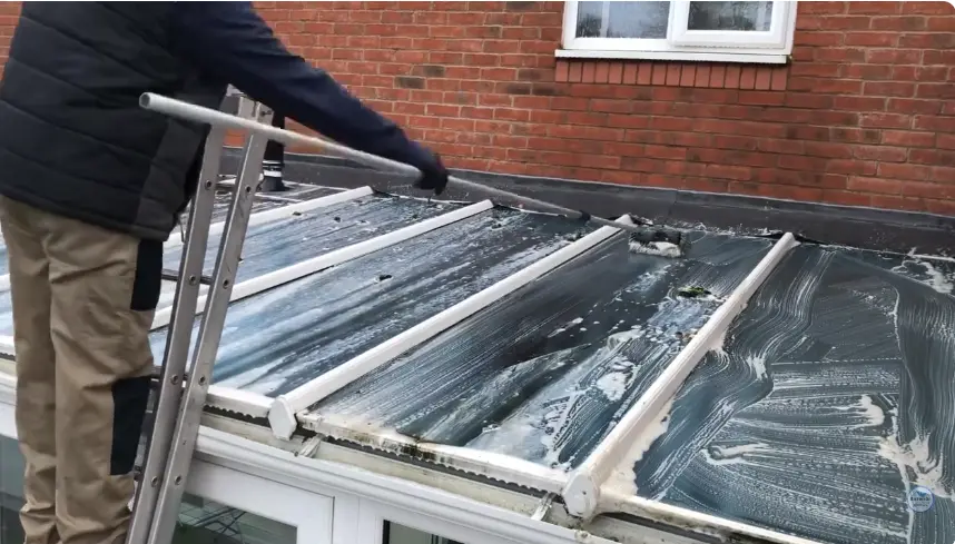 Cleaning Glass Roof Panels From a Ladder Technician on a ladder cleaning sloped glass roof panels beside a brick wall