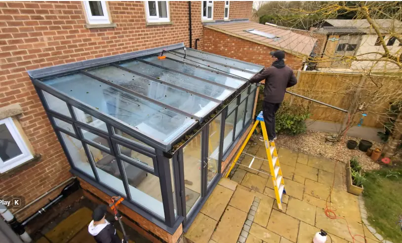 Cleaning a Glass Roof From a Ladder Window cleaner on a ladder beside a glass-roofed sunroom on a brick house