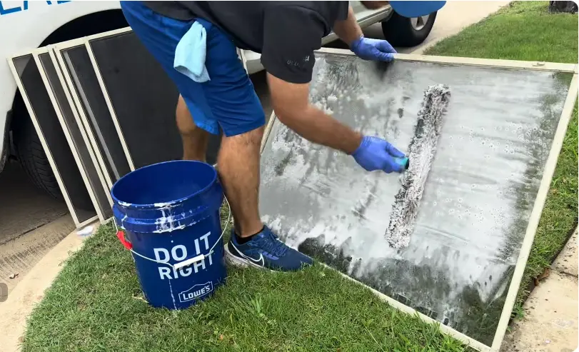Washing a Window Screen With Soapy Solution Window screen covered in soapy cleaning solution being scrubbed next to a bucket, with more screens waiting