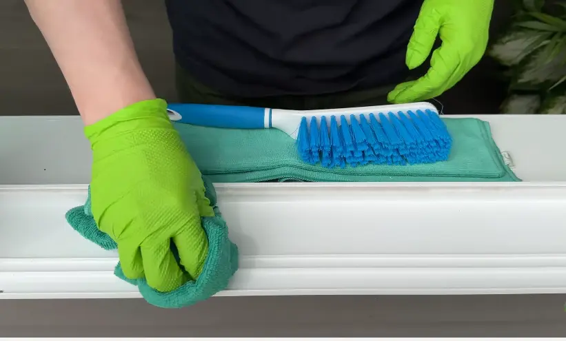 Window Sill Cleaning With a Microfiber Cloth Gloved hands wiping a white window sill with a microfiber cloth beside a scrub brush